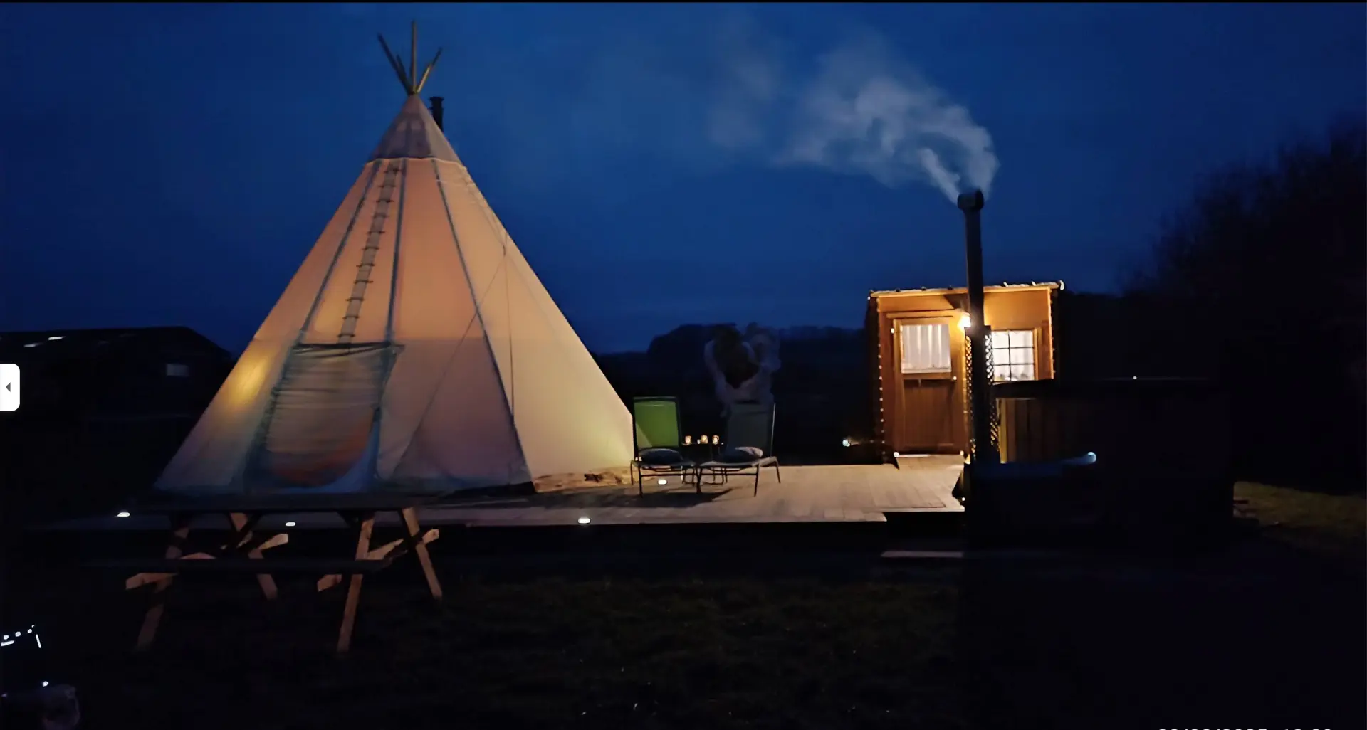 Le tipi et le chalet sanitaire éclairés à la tombée de la nuit, fumée s’échappant du poêle du bain nordique
