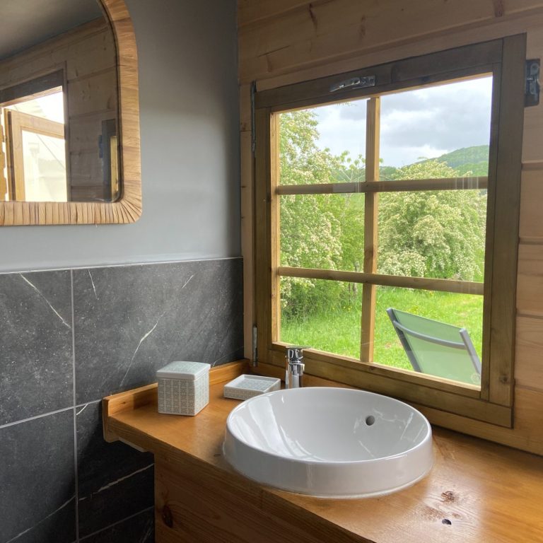 Salle d’eau privative en bois avec vasque ronde, miroir et fenêtre ouvrant sur la campagne